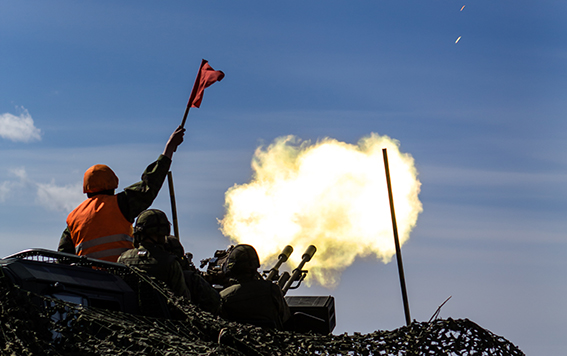 Sotilas ampuu ilmatorjuntatykillä, vieressä oranssiliivinen sotilas pitää punaista lippua ilmassa.