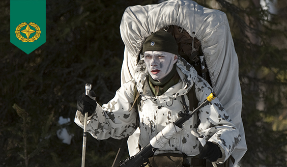 A soldier is skiing, carrying a big backpack, in a white snow suit, his face painted white