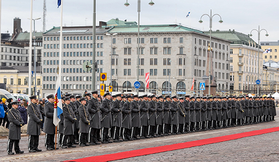 Soldiers stand in a row in the city wearing gray clothes. In front of them is a long red carpet.