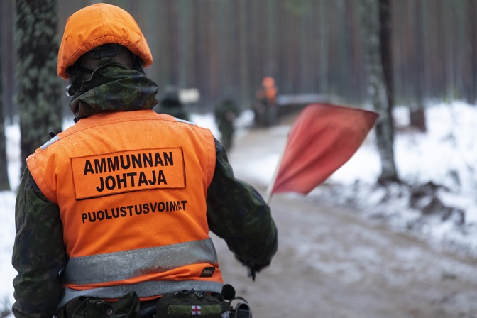 Sotilaskouluttaja oranssissa liivissä ja kypärässä punainen lippu kädessä, maastopuvussa. Ammunnanjohtaja käsikranaatin heiton harjoituksessa.