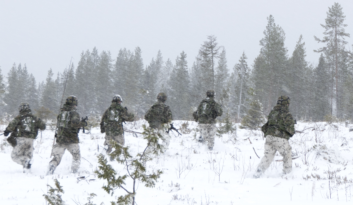 Kuusi varusmiestä etenee juosten metsässä, lunta tuiskuttaa sakeasti.
