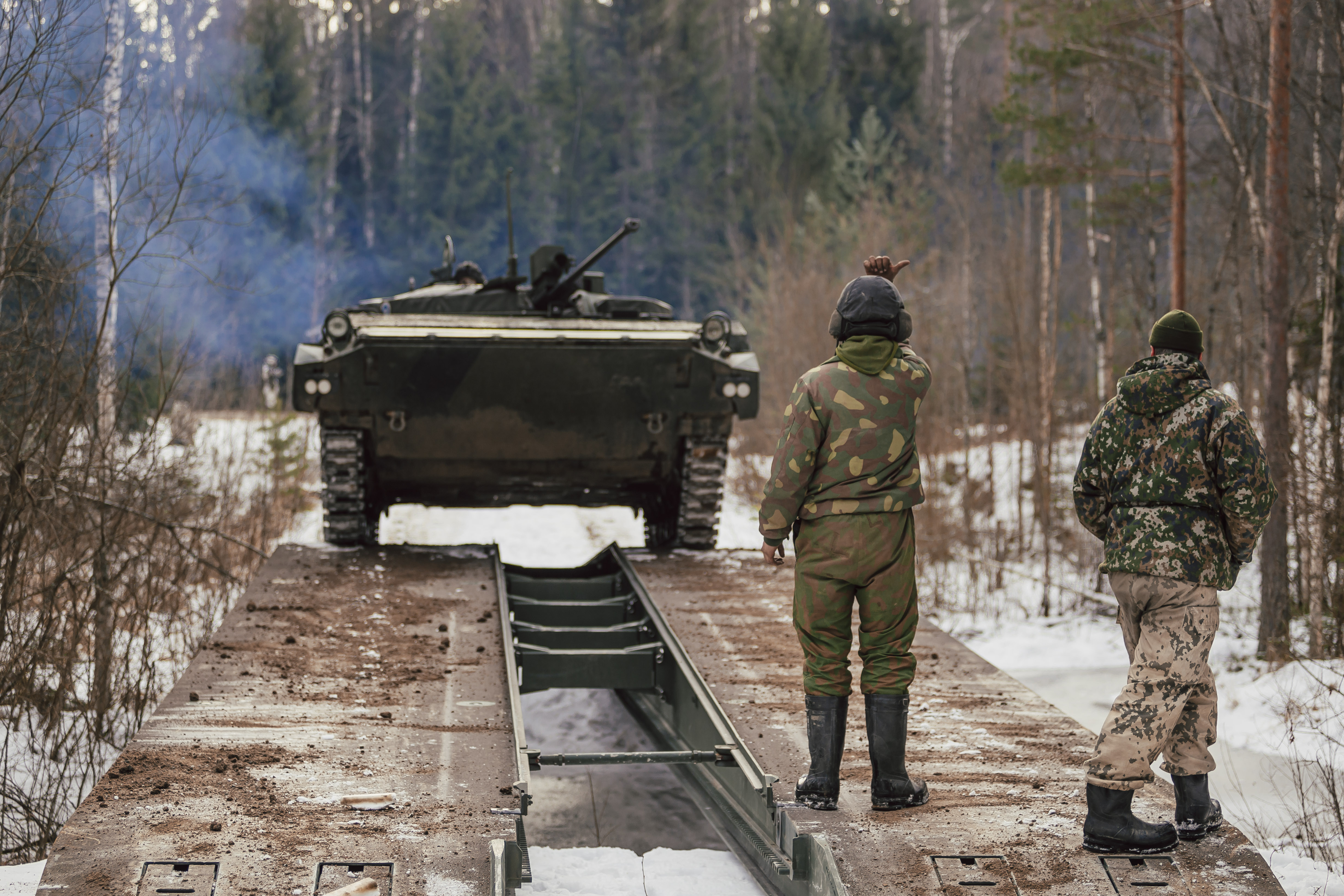 Panssarivaunu siirtymässä perustetulla sillalla yli joen, panssarirukkilainen ohjaa tulemaan ja kouluttaja valvoo.