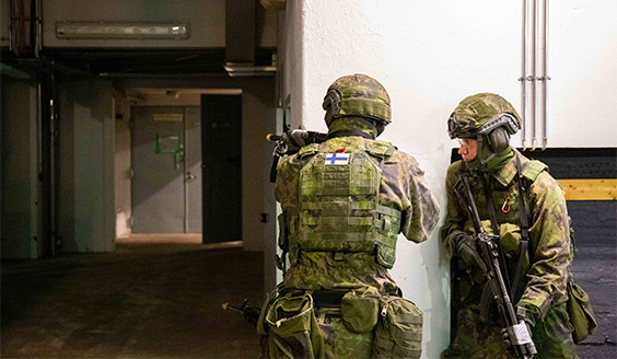 Kuvassa kaksi sotilaspoliisia taisteluvarusteissa rakennuksen sisällä. Toinen osoittaa aseella käytävän päässä kulman takana ja toinen hänen takanaan.