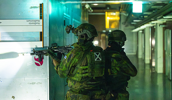 Kuvassa kaksi sotilasasuista henkilöä käsissään aseet seisomassa käytävällä.