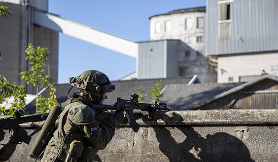 Kuvassa on sotilas rynnäkkökiväärin kanssa ja taustalla tehdasrakennus ja tehtaan piippu.