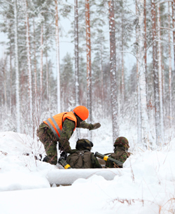 A trainer in an orange vest is pointing to the forest while two soldiers listen and observe