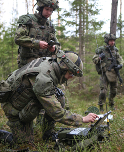 Soldiers in the forest in combat equipment. One is using a computer.