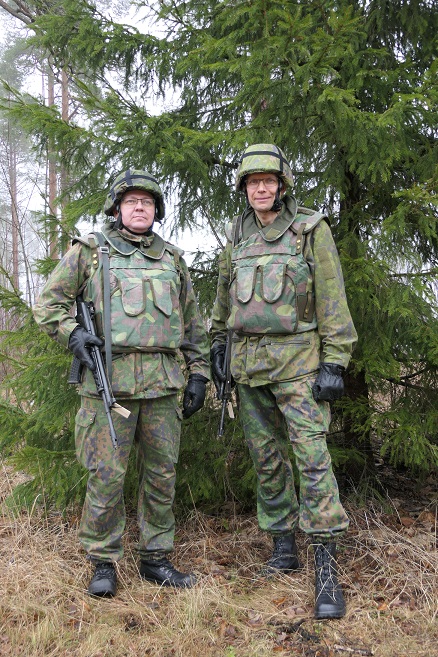 Kaksi maastopukuista miestä seisoo aseistettuna kuusen vieressä.