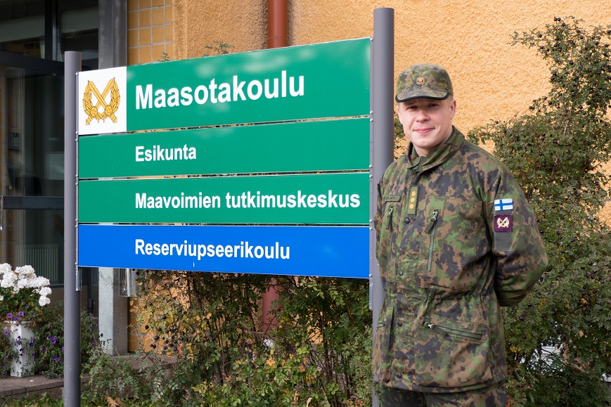 Kapteeni Antti Paronen maastopuvussa seisoo Maavoimien tutkimuskeskuksen ja RUK:n rakennuksen edessä.
