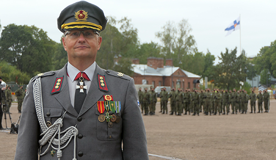eversti Pietiläinen taustallaan varusmiehiä, rakennuksia ja Suomen  lippu