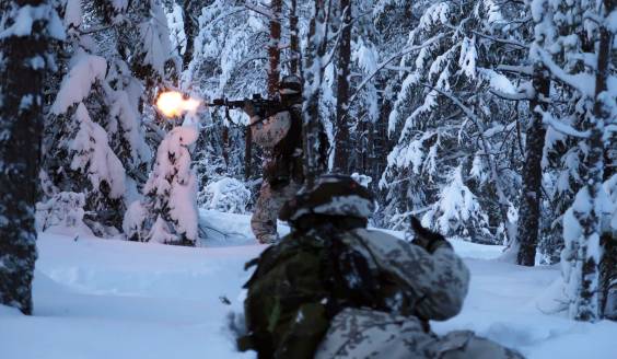 sotilaat harjoittelevat talvella metsässä, toinen on maassa ja toinen ampuu juostessaan