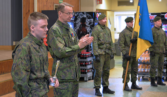 Porin prikaatin komentaja taputtaa vieressään seisovalle urheilijapalkinnon saaneelle varusmiehelle Niinisalon sotilaskodissa. Taustalla prikaatin lippusaattue.