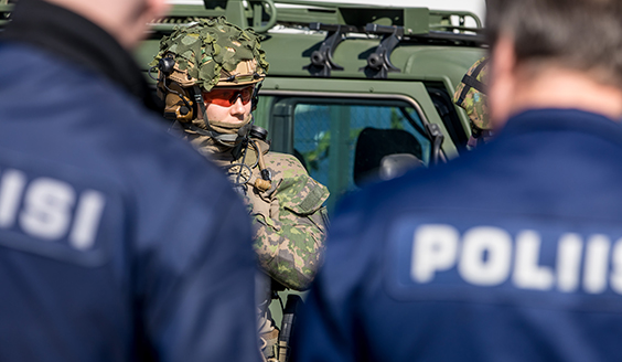 Kuvassa kaksi poliisia selkä vasten kameraa sekä taisteluvarustuksessa oleva reserviläinen kasvot kohti kameraa. Taustalla puolustusvoimien ajoneuvo.