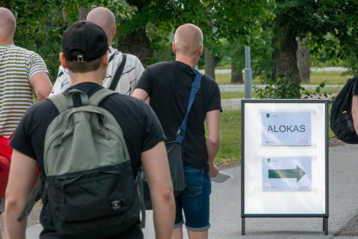 Kuvassa kaksi tuoretta alokasta siviilivaatteissa ja heidän vieressään on kyltti, jossa lukee alokas ja nuoli oikealle.
