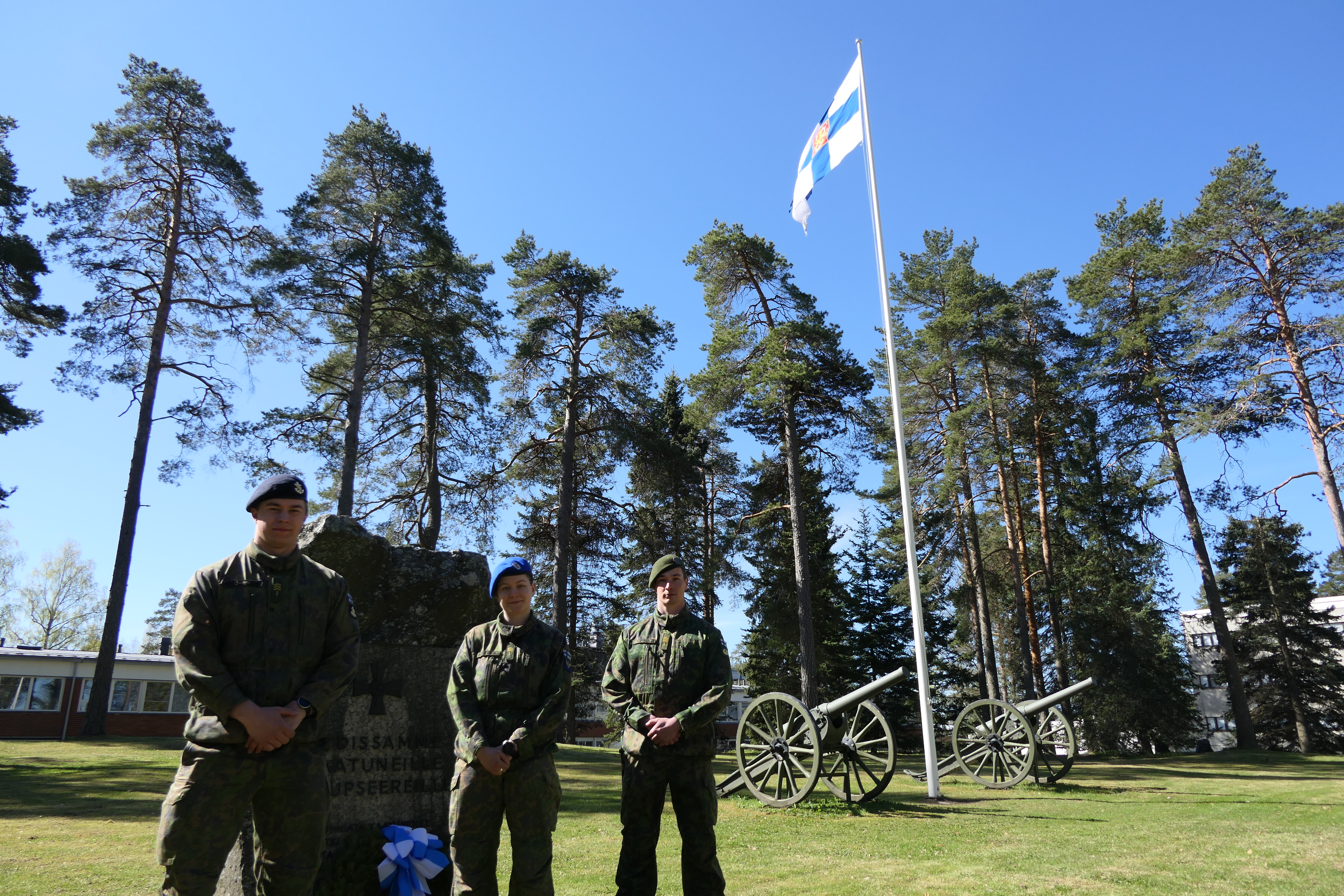 Puolustusvoimain lippu liehuu taustalla, edessä seisoo kolme maastopukuista sotilasta. 