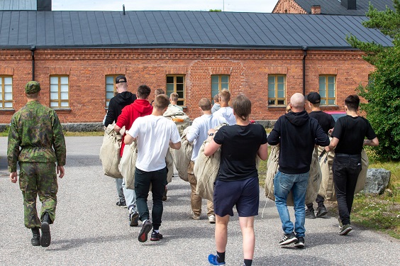 Alokkaat kantavat siviilivaatteissaan suuria varustesäkkejä varusvarastolta kasarmille varusmiesjohtajan seuratessa mukana.