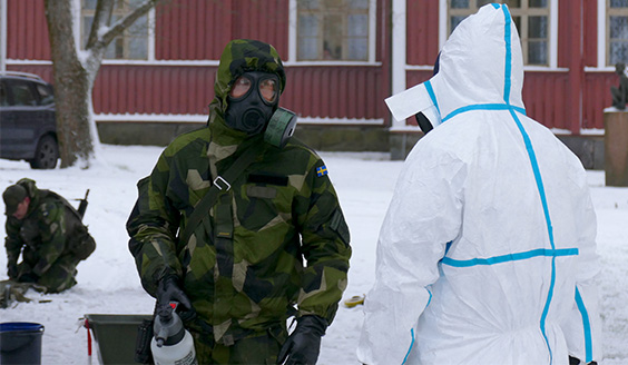 Ruotsalainen sotilas suojanaamari päässä ja painesuihkupullo kädessä katsoo valkoisessa suojapuvussa olevaa henkilöä.