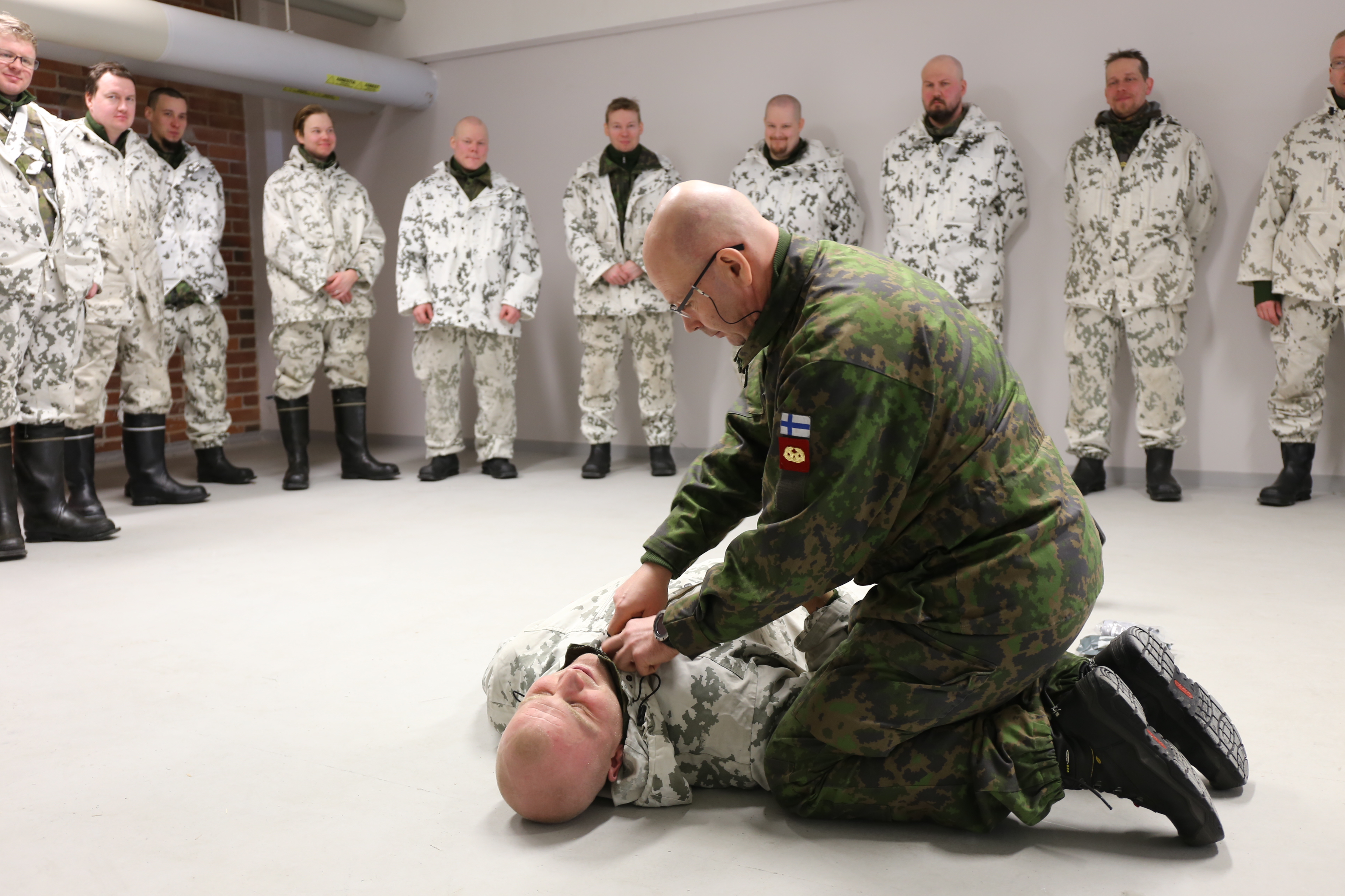 Lumipukuiset sotilaat seuraavat koulutusta jossa mies makaa maassa ja kouluttaja pitää kiinni hänen kauluksestaan