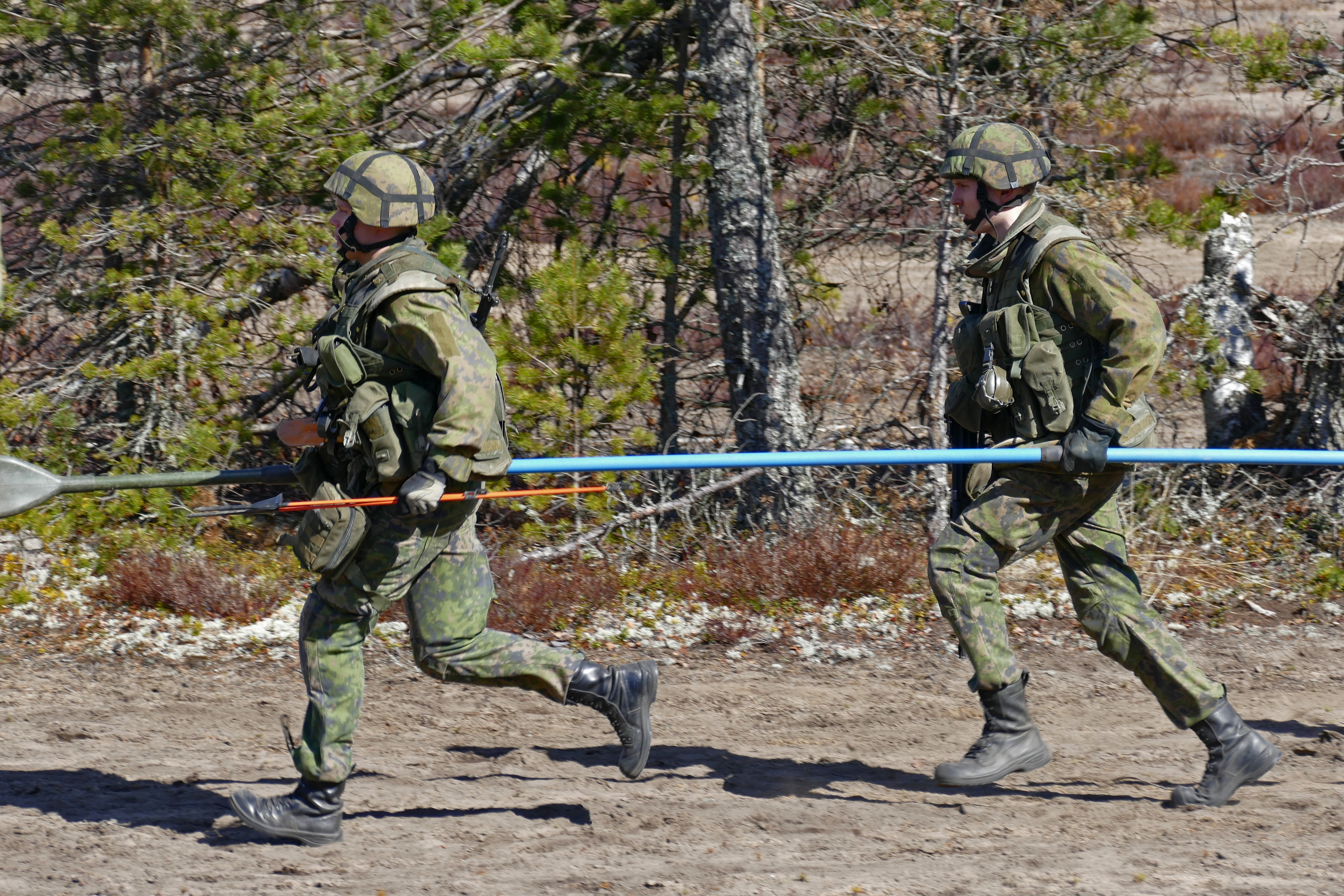 Kaksi sotilasta juoksee metsätietä pitkin kantaen sinistä keppiä