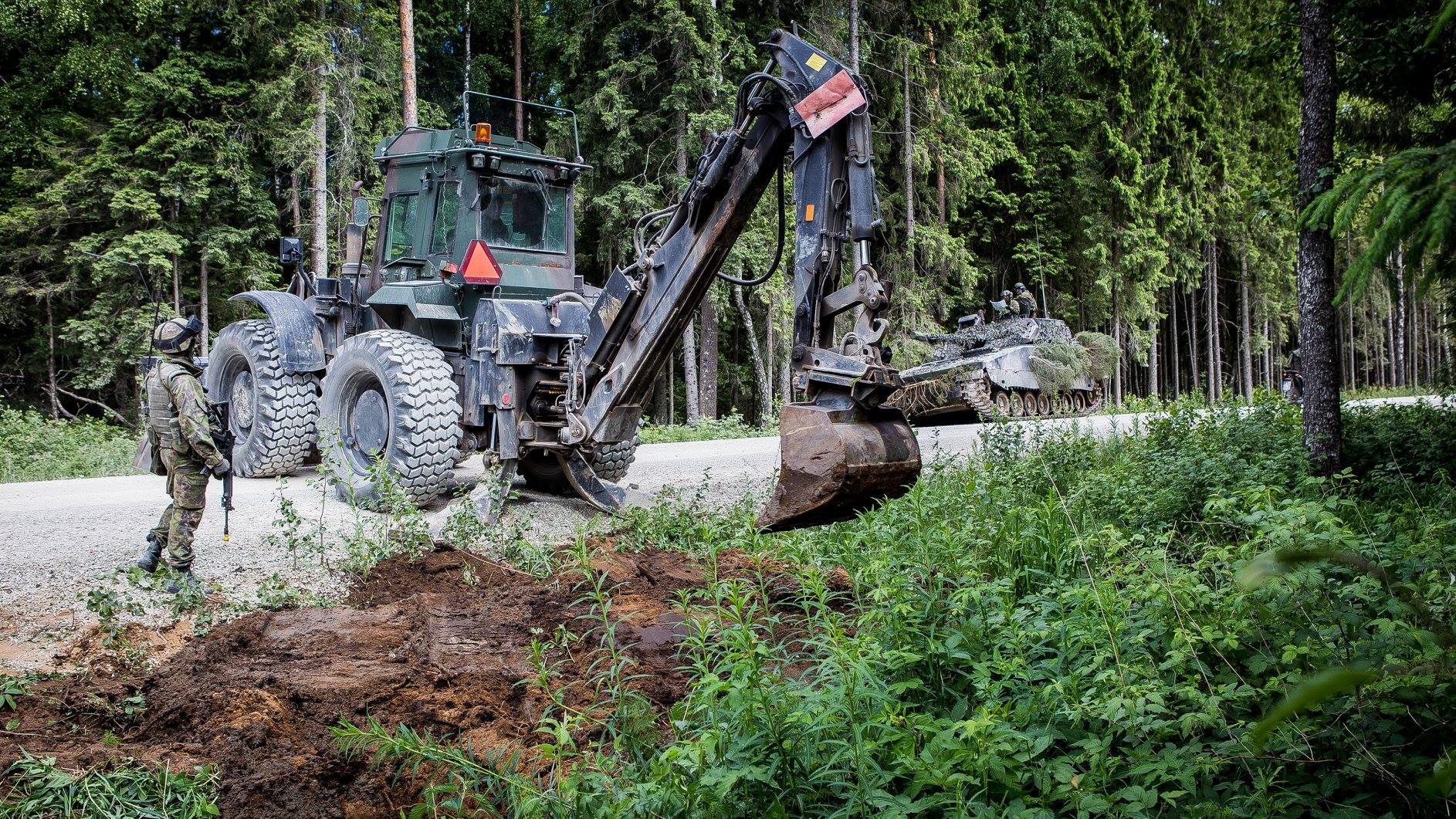 Kaivinkone kaivaa piennarta hiekkatiellä ja sotilas seuraa vierestä