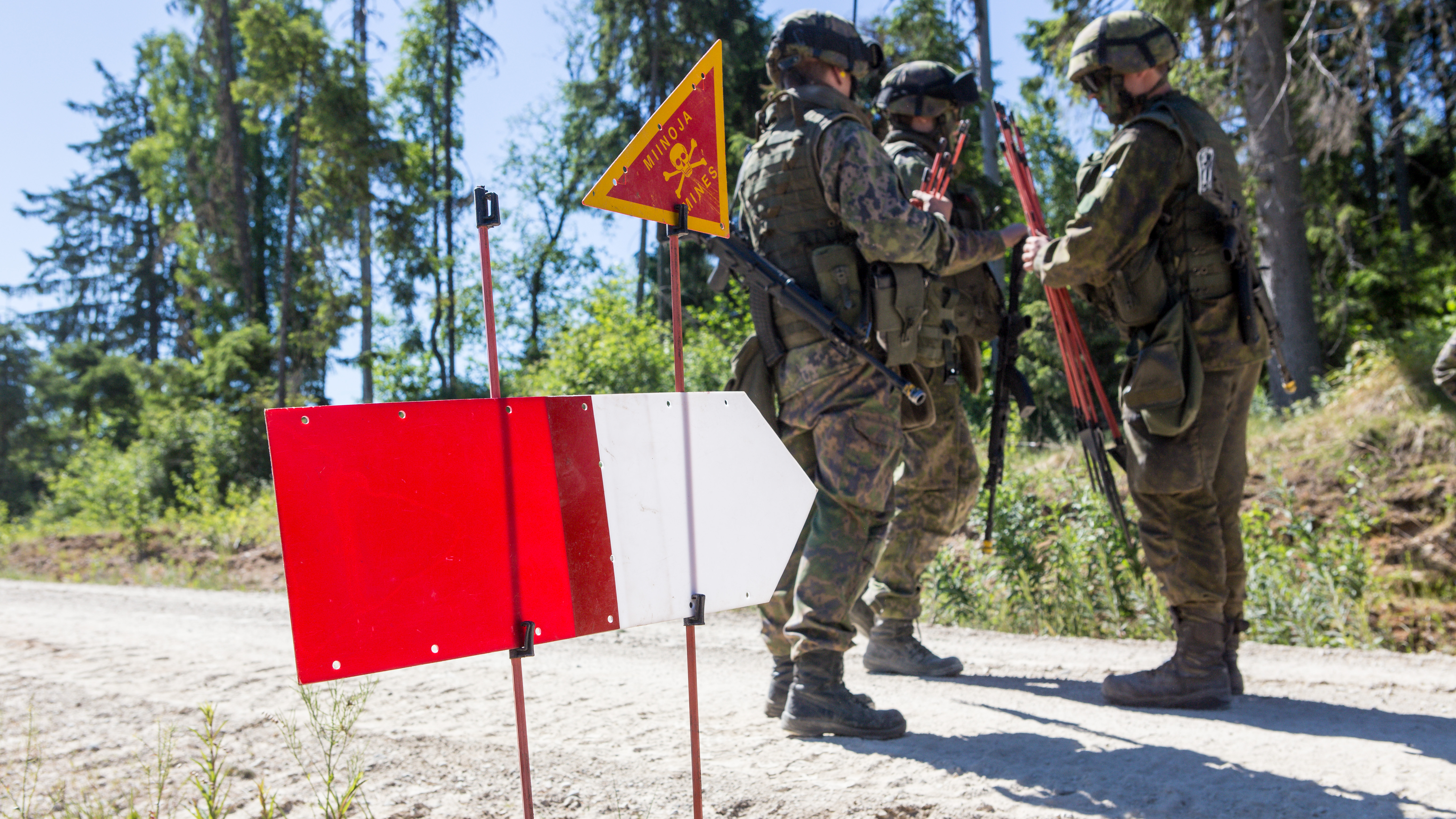 Soldiers standing on a dirt road with red canes in hand. Along the road is a sign that says "Miinoja, Mines."