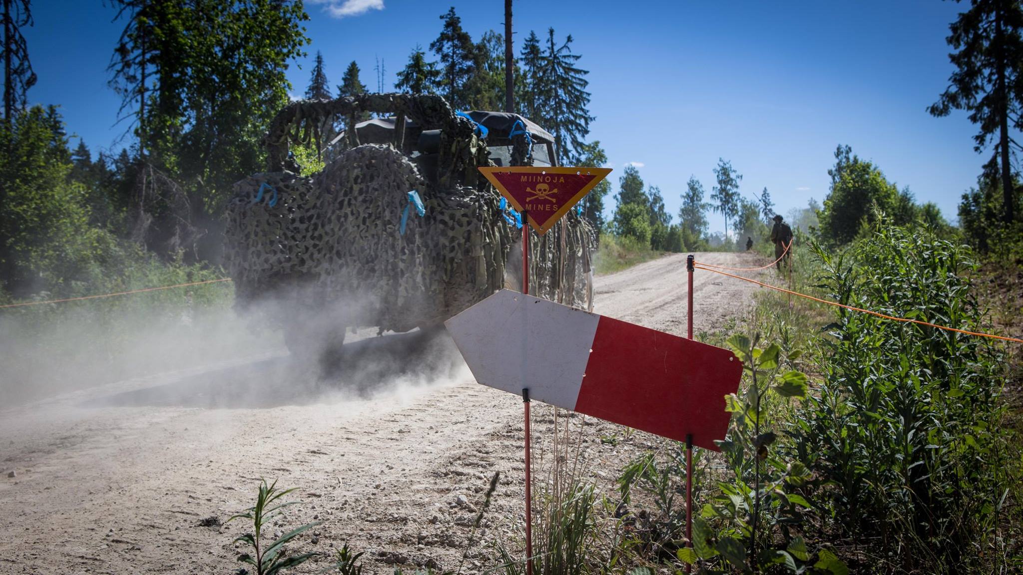 Naamioitu ajoneuvo ajaa metsätietä, jonka reunassa kyltti jossa lukee "miinoja, mines"