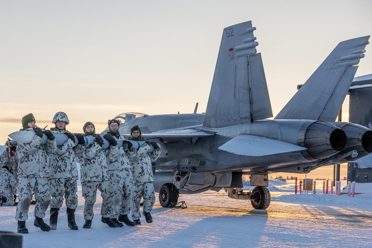Viisi lumipukuista varusmiestä kantaa AMRAAM-ohjusta sylissään. Taustalla lumisella alustalla seisoo Hornet-hävittäjä.