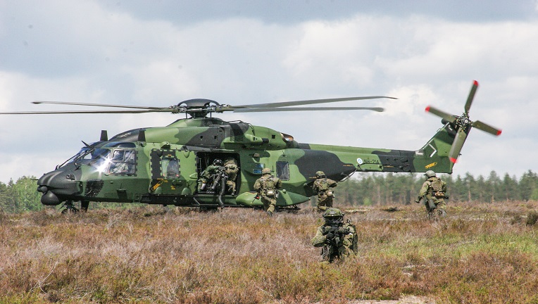 NH90-helikopteri laskeutuneena niitylle. Helikopterin edessä liikkuu neljä aseistautunutta sotilasta.