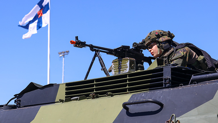 Varusmies tähtää PKM-konekiväärillä panssaroidun ajoneuvon katolta. Aurinkoinen sää ja Suomen lippu liehuu taustalla.